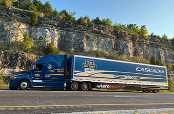 A blue Freightliner Cascadia semi-truck with a trailer drives along a rocky highway.